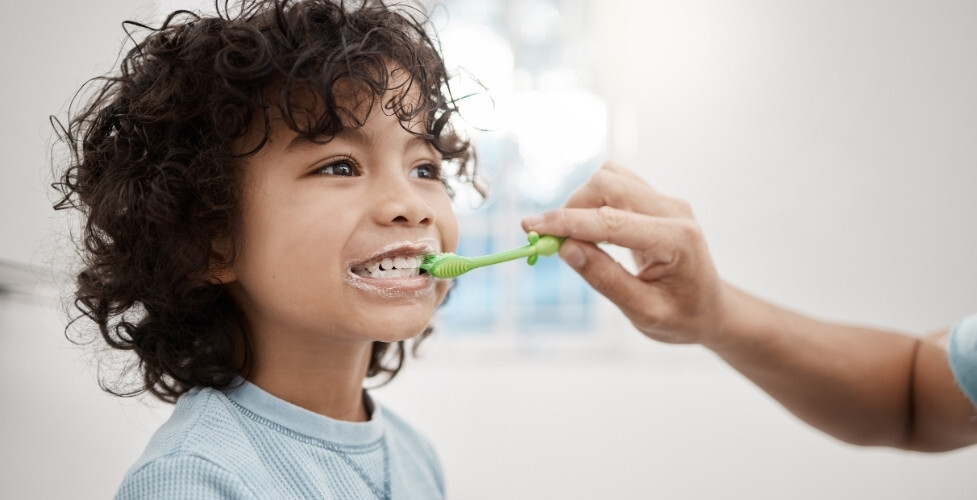 Young child with curly dark hair brushing teeth with a green toothbrush during preventive dental care.