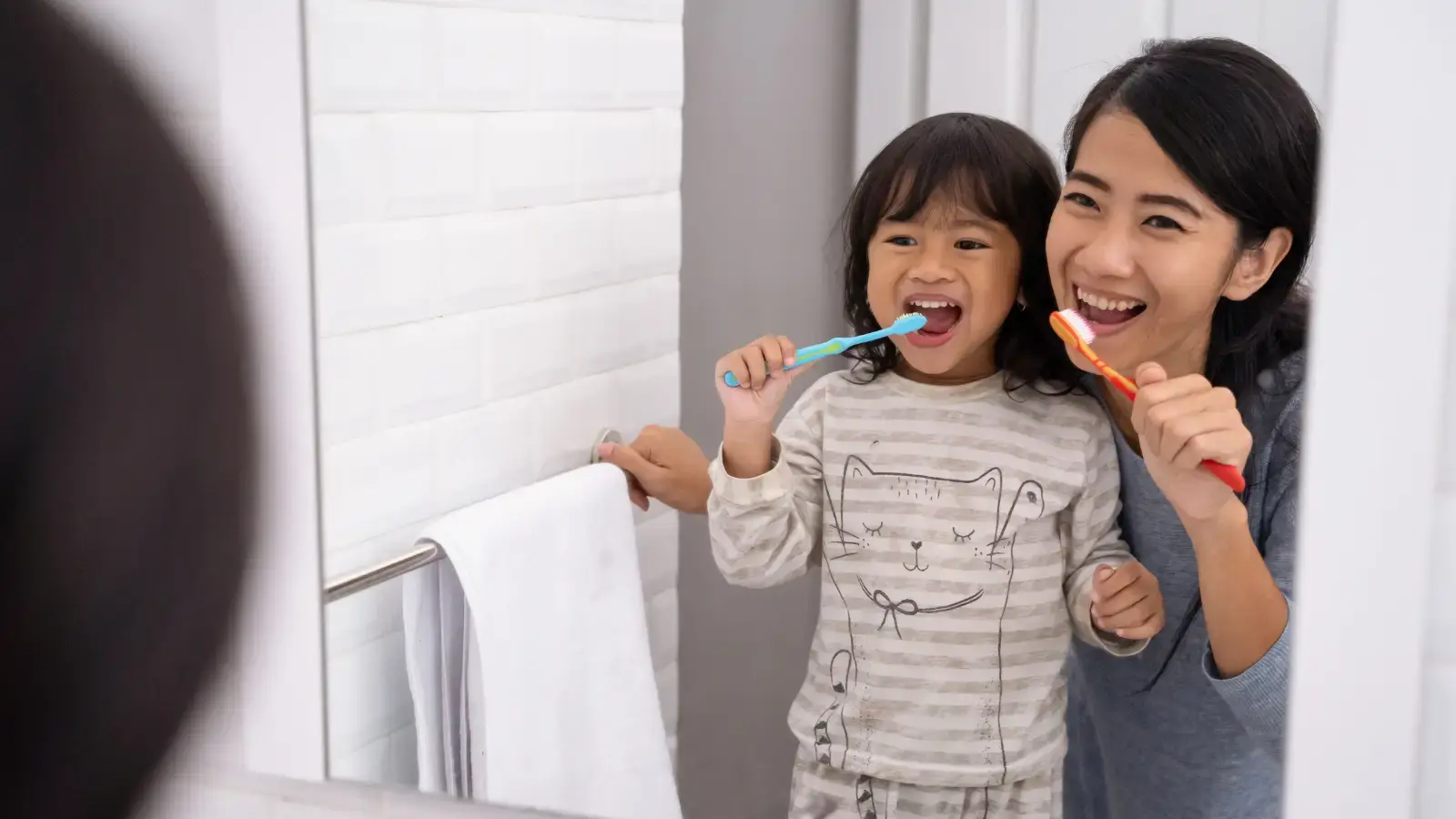 Mother and daughter brushing teeth together in bathroom, smiling while practicing good oral hygiene habits.