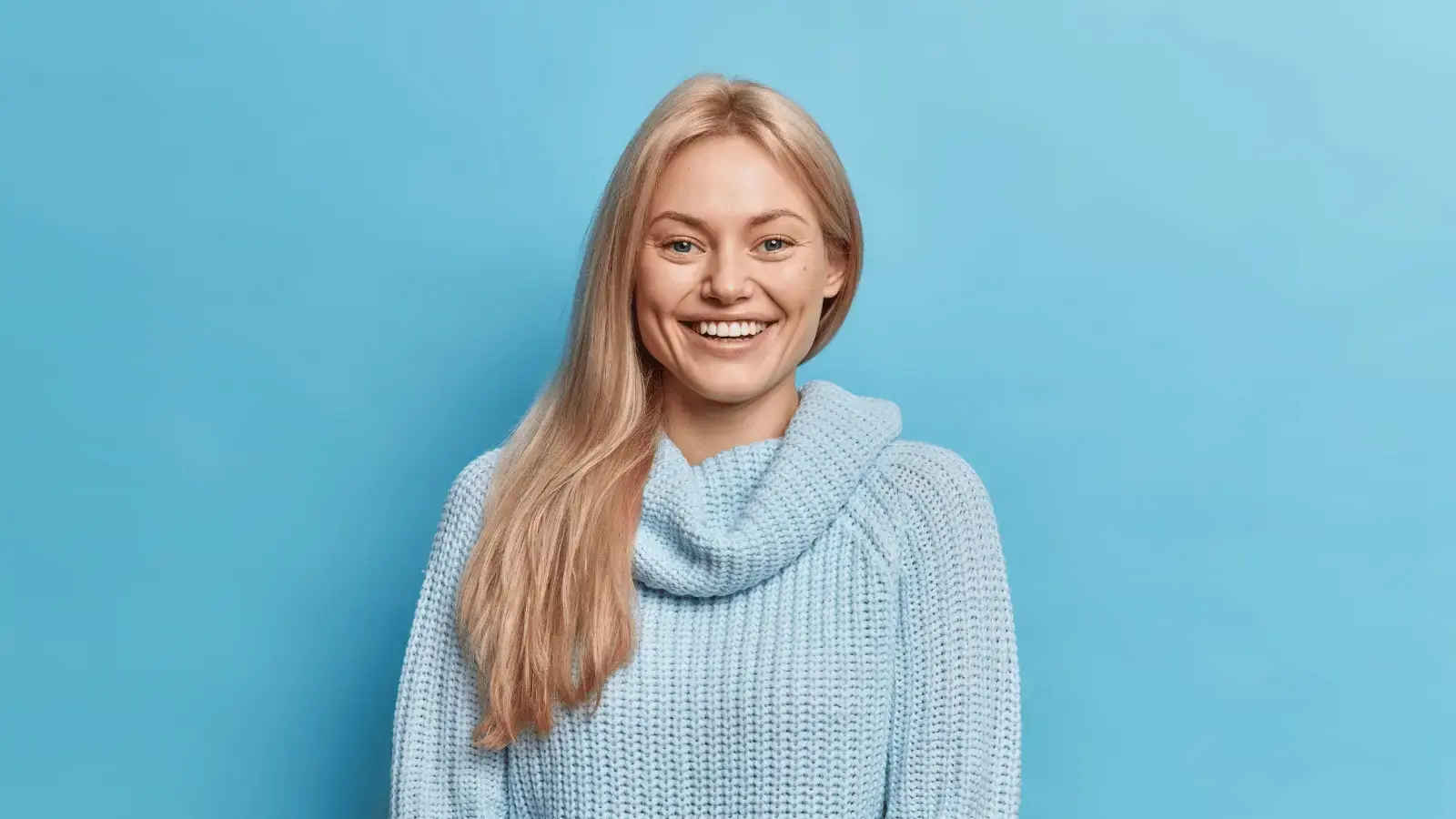 Smiling woman with bright teeth wearing light blue sweater against blue background.