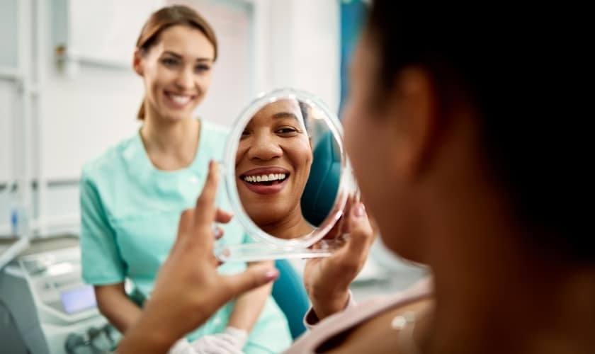 Female patient smiling at mirror while dental hygienist in teal scrubs watches approvingly in dental office.