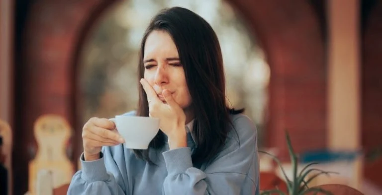 Woman wincing and holding her cheek while drinking from a cup